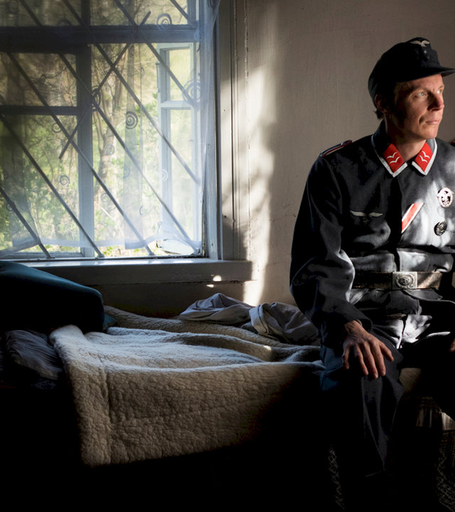 "Der Krieg in mir": Sebastian Heinzel sitzt in Wehrmachtsuniform auf einem Einzelbett im Halbdunkel und schaut nachdenklich. Im Hintergrund sieht man Bäume durch das geöffnete Fenster.