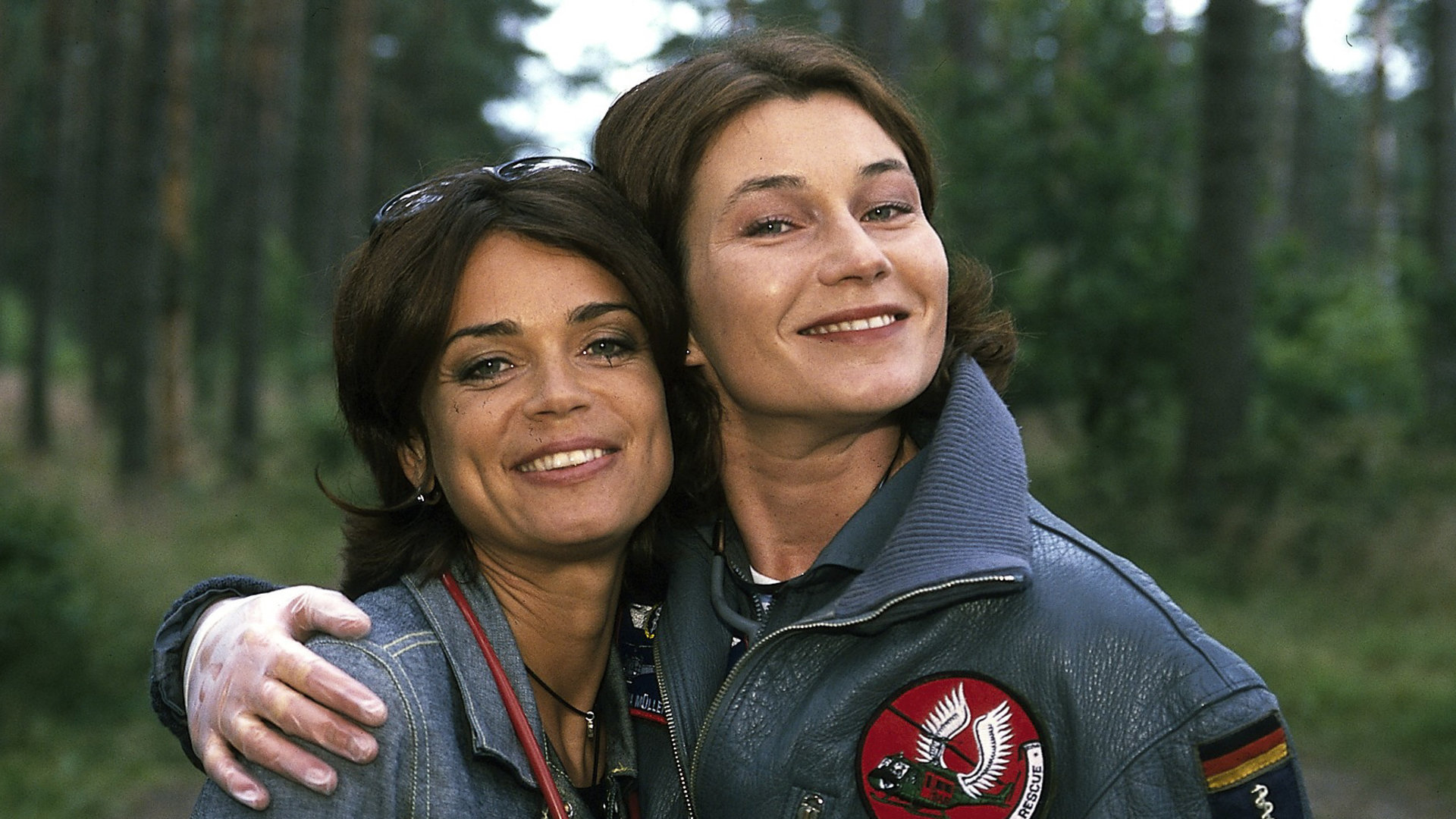 "Die Rettungsflieger - Abschied von Maren": Außenaufnahme im Wald - Maren (Gerit Kling) und Ilona (Julia Heinemann) stehen eng beieinander und strahlen in die Kamera.