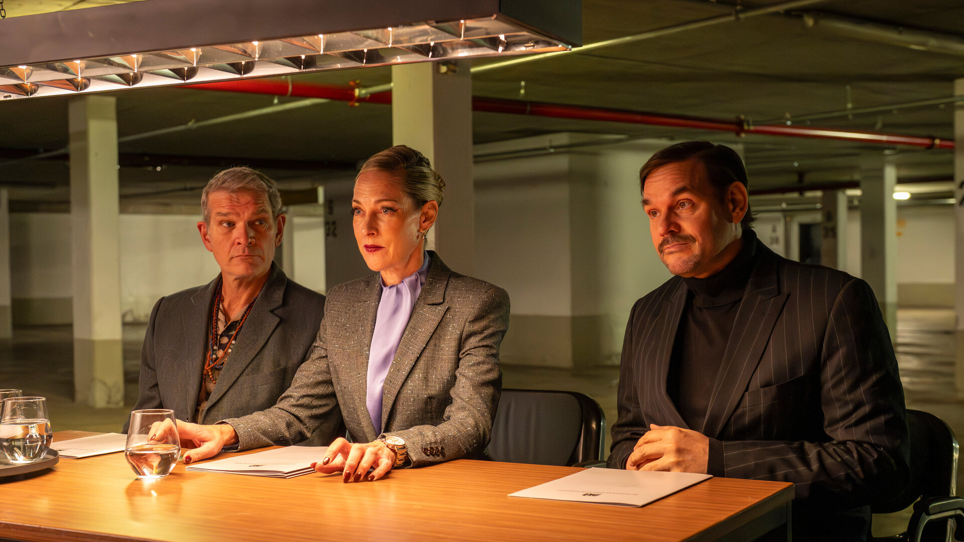 "SPYON - Der perfekte Spion": Ausbilder Voss (Götz Otto), Ausbilderin Haberland (Sophie von Kessel) und Ausbilder Kocabay (Alexander Beyer) sitzen nebeneinander an einem Tisch und schauen in die Ferne. Vor den dreien liegen Bewerbungsmappen.