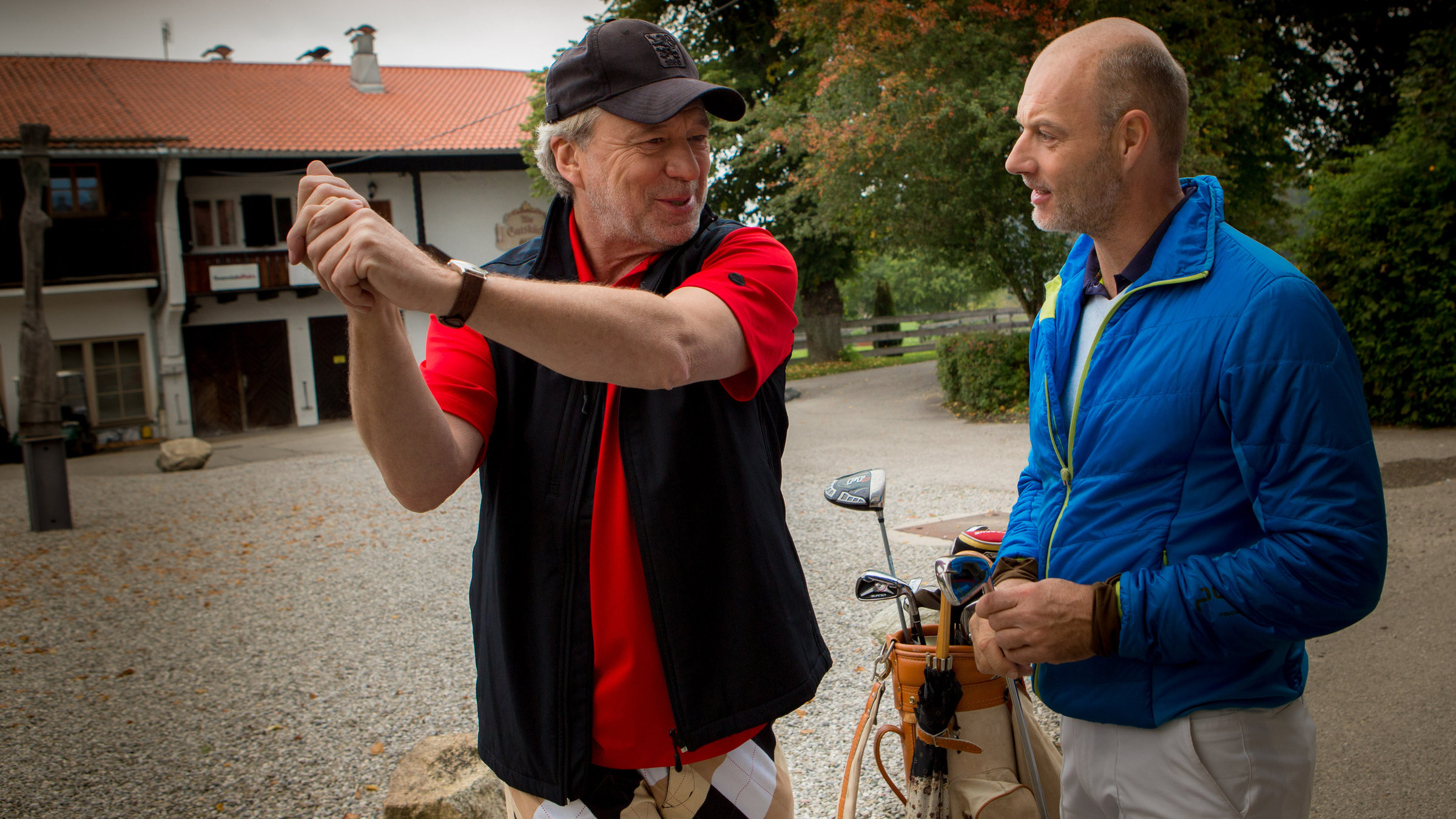 "SOKO München - Der Golfsack": Arthur Bauer (Gerd Silberbauer) und Dean Bradley (Simon Licht) unterhalten sich über Golf. Bauer macht eine Abschlagbewegung. Bradley hält einen Golfschläger in der Hand, zwischen den beiden steht eine Golftasche, im Hintergrund ist der Golfclub zu sehen.