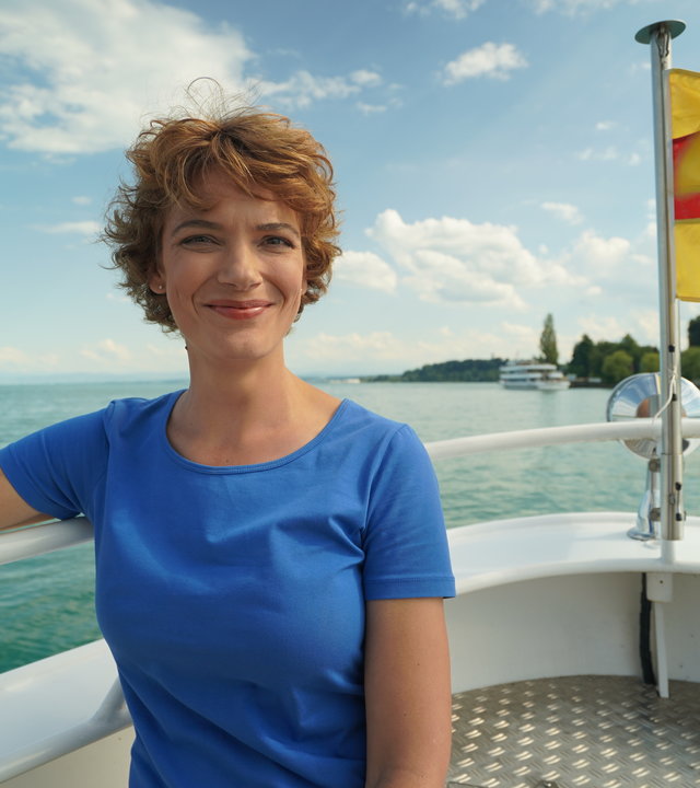"Terra Xpress: Faszination Bodensee - Leben am Wasser": Moderatorin Lena Ganschow sitzt in der Spitze eines Bootes: Im Hintergrund weht eine gelb-rote Fahne und man sieht in der Ferne Land.