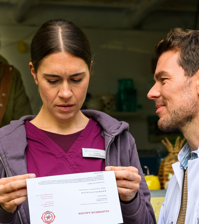 "Bettys Diagnose - Yesterday": Betty (Henrike Hahn) hält einen Brief in beiden Händen und Dr. Walden (Constantin Lücke) schaut sie dabei an. Im Hintergrund beobachtet Safi (Ercan Durmaz) die beiden aus seinem Imbisswagen heraus.