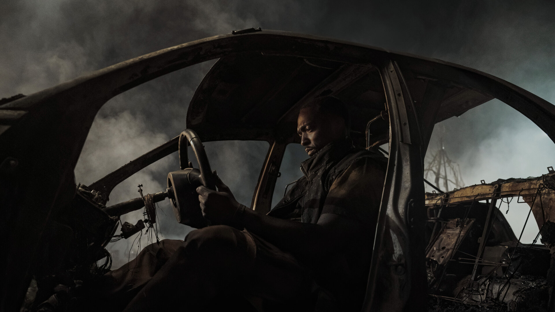 "Twisted Metal - EV3L1N": John (Anthony Mackie) sitzt nachdenklich in einem ausgebrannten Autowrack und hält das Lenkrad in den Händen. Dichte Rauchschwaden umgeben das Fahrzeug.