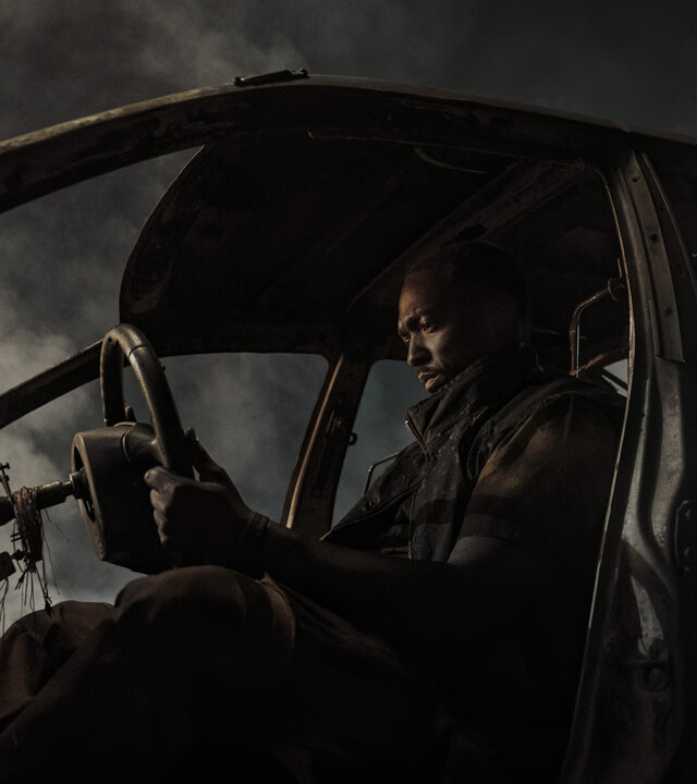 "Twisted Metal - EV3L1N": John (Anthony Mackie) sitzt nachdenklich in einem ausgebrannten Autowrack und hält das Lenkrad in den Händen. Dichte Rauchschwaden umgeben das Fahrzeug.