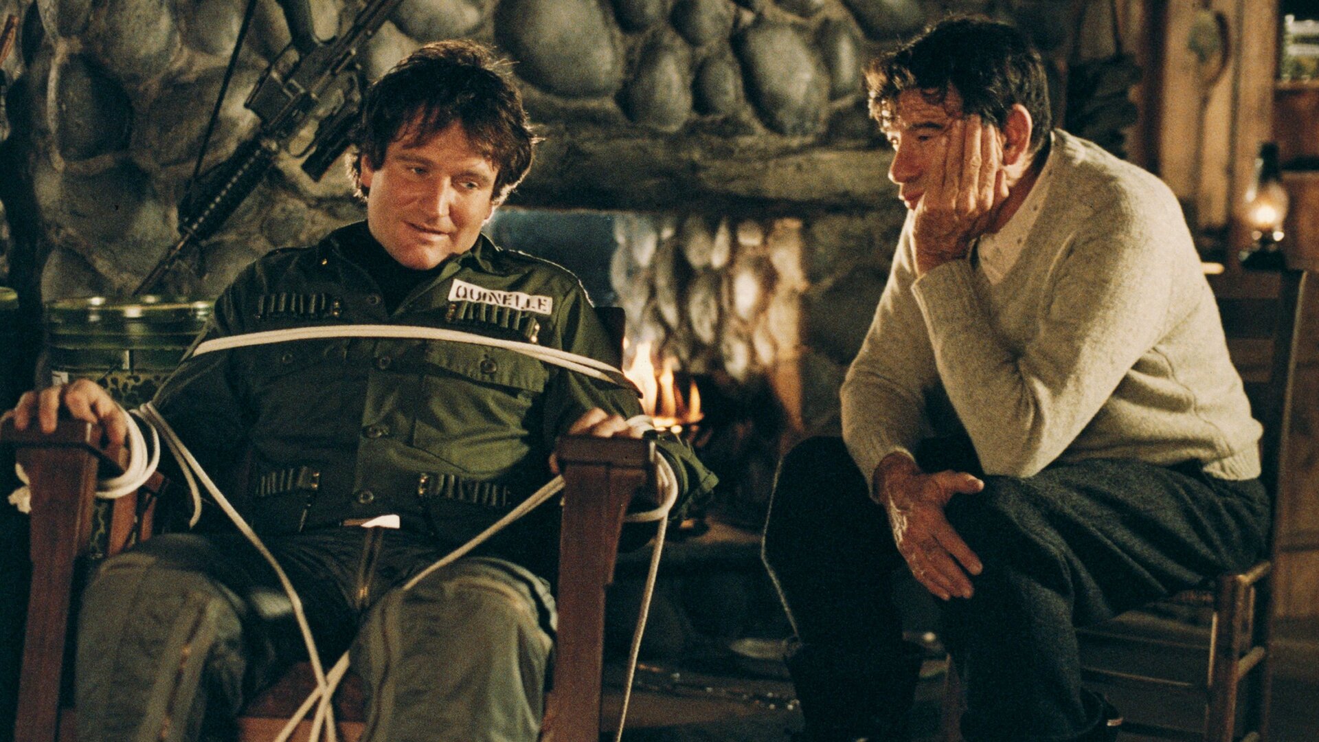 "Die Überlebenskünstler": Donald Quinelle (Robin Williams) sitzt gefesselt auf einem Stuhl. Sonny Paluso (Walter Matthau) sitzt daneben und betrachtet ihn ruhig. Er stützt sein Kinn auf einer Hand ab.