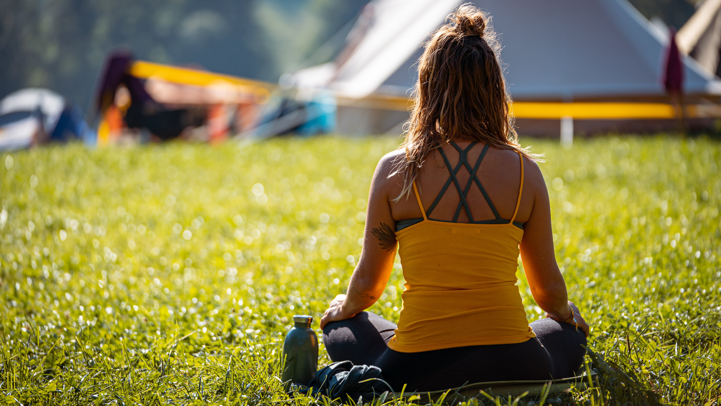 "Rituale, Esoterik, Aberglaube: Sinnsuche zwischen Spiritualität und Wissenschaft": Frau von hinten im Schneidersitz auf Wiese vor Zelten.