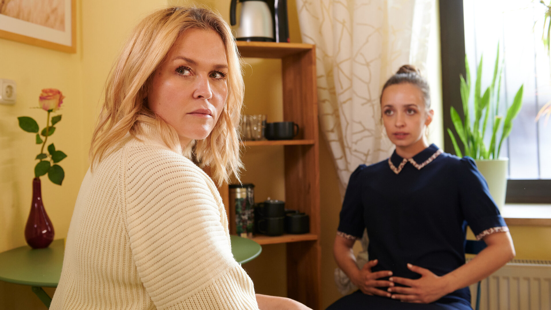"Lena Lorenz - Lügenbaby": Lena Lorenz (Judith Hoersch) sitzt Flora Bergmann (Nellie Thalbach) in einem Aufenthaltsraum gegenüber. Flora hält sich ihren schwangeren Bauch, Lena hat sich nach vorne gedreht und blickt ernst.