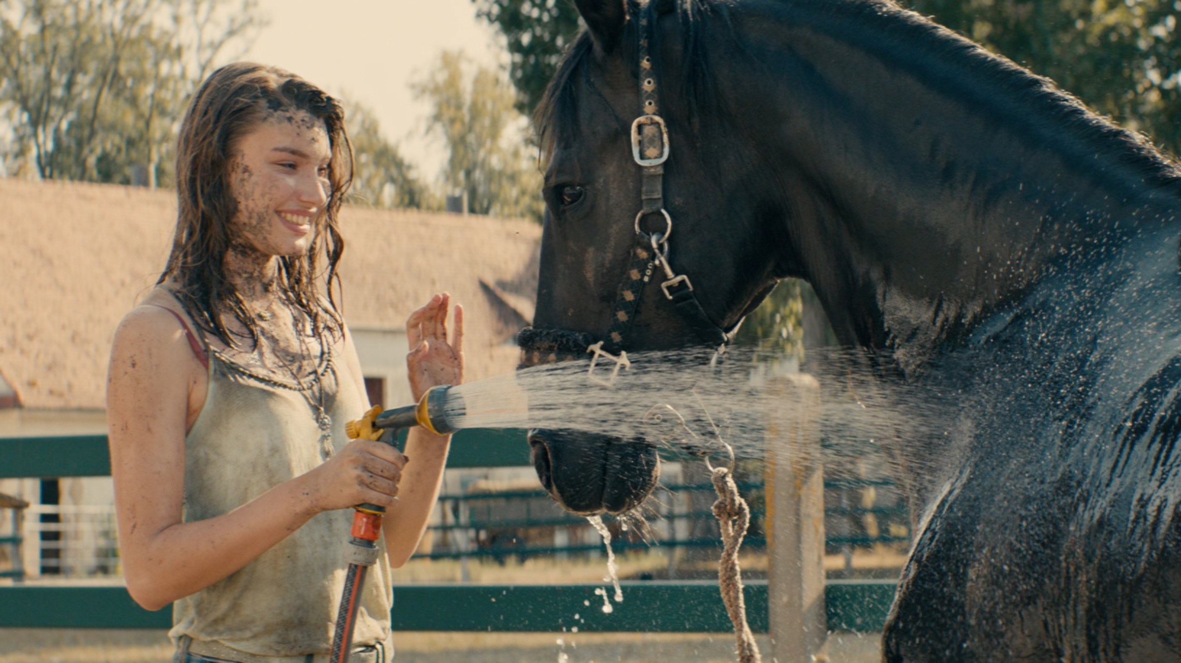 "Immenhof - Das Abenteuer eines Sommers": Lou (Leia Holtwick) steht mit schmutzigem Gesicht lachend vor einem Rappen und spritzt ihn nass.