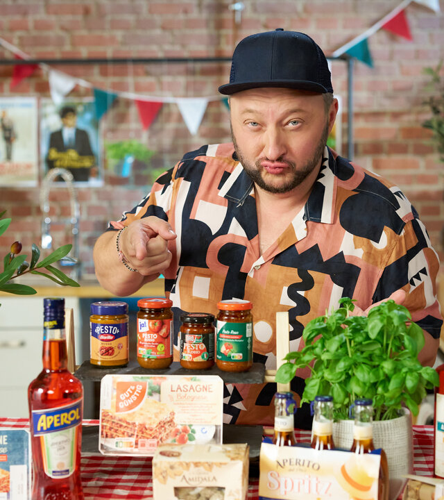 "besseresser: Italienische Imitate - Sebastian Lege packt aus": Sebastian Lege steht hinter einem Tisch mit italienischen Produkten und zeigt auf diese.