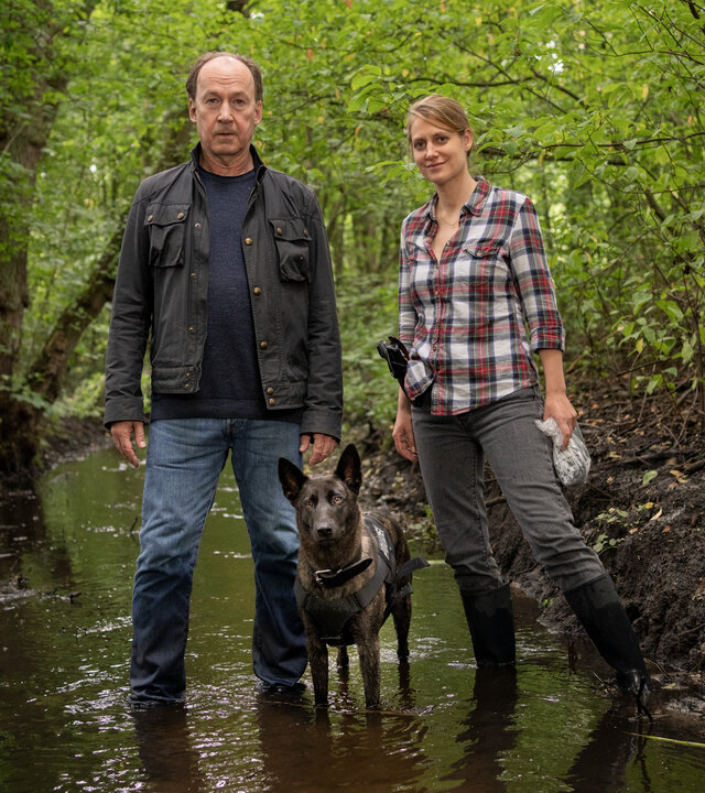 "Wendland - Stiller und der Wolf im Schafspelz": Jakob Stiller (Ulrich Noethen) und Kira Engelmann (Bettina Burchard) stehen zusammen mit einem Hund im Wald in einem Bach.