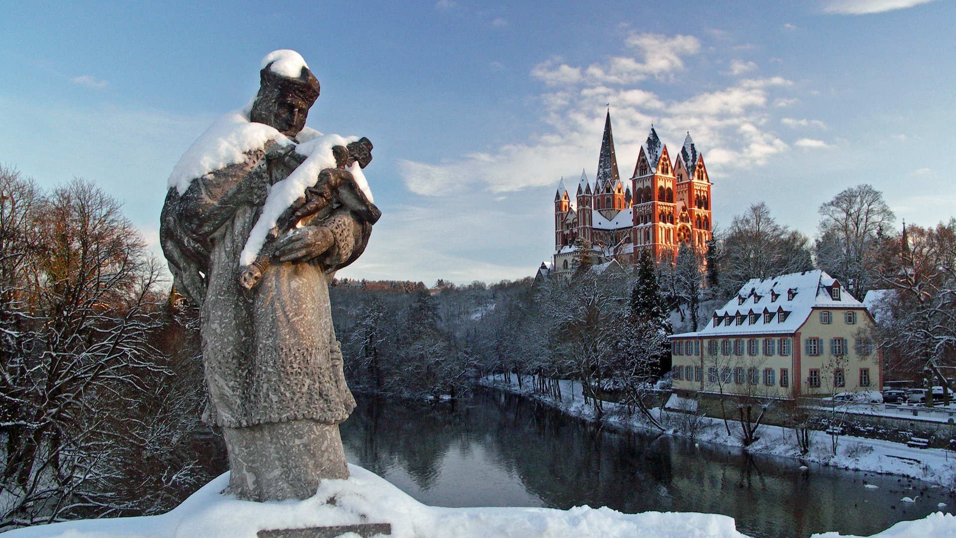"Katholischer Weihnachtsgottesdienst - Ehrenwort!":  In einer Winterlandschaft steht im Vordergrund die Statue des Heiligen Johann von Nepomuk, dem Schutzpatron der Brücken, auf einer Brücke über die Lahn. Dahinter liegt der Limburger Dom im Sonnenschein.
