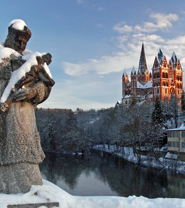 "Katholischer Weihnachtsgottesdienst - Ehrenwort!":  In einer Winterlandschaft steht im Vordergrund die Statue des Heiligen Johann von Nepomuk, dem Schutzpatron der Brücken, auf einer Brücke über die Lahn. Dahinter liegt der Limburger Dom im Sonnenschein.