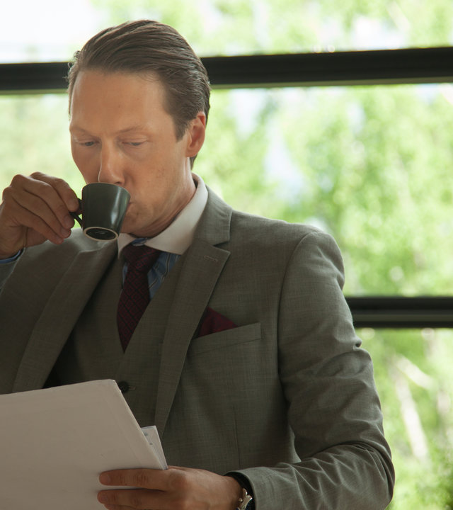 "Exit - Macht": Adam Veile (Simon J. Berger) hält sich mit der rechten Hand eine Espresso-Tasse an die Lippen und blickt gleichzeitig nach unten auf ein Blatt Papier in seiner linken Hand. Adam trägt einen grauen Anzug.