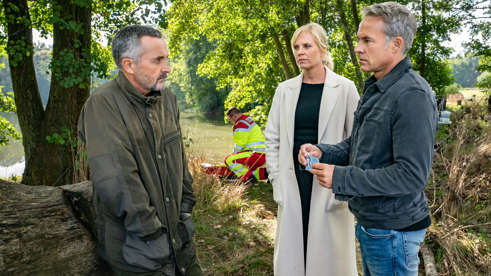 "SOKO Leipzig - Family Business (1)": Davide Di Maria (Harald Schrott) steht Ina Zimmermann (Melanie Marschke) und Jan Maybach (Marco Girnth) am Ufer eines Sees gegenüber. Jan zeigt Davide seinen Dienstausweis. Im Hintergrund kniet ein Sanitäter (Komparse) vor einer Leiche.