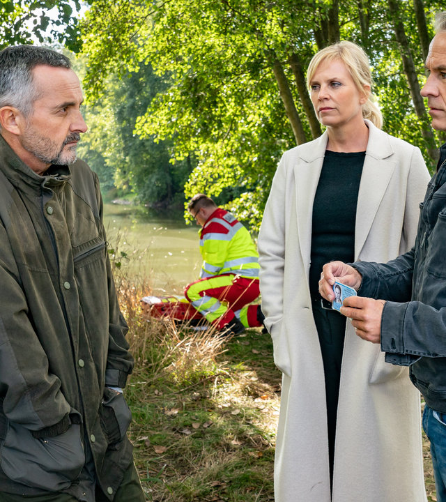 "SOKO Leipzig - Family Business (1)": Davide Di Maria (Harald Schrott) steht Ina Zimmermann (Melanie Marschke) und Jan Maybach (Marco Girnth) am Ufer eines Sees gegenüber. Jan zeigt Davide seinen Dienstausweis. Im Hintergrund kniet ein Sanitäter (Komparse) vor einer Leiche.