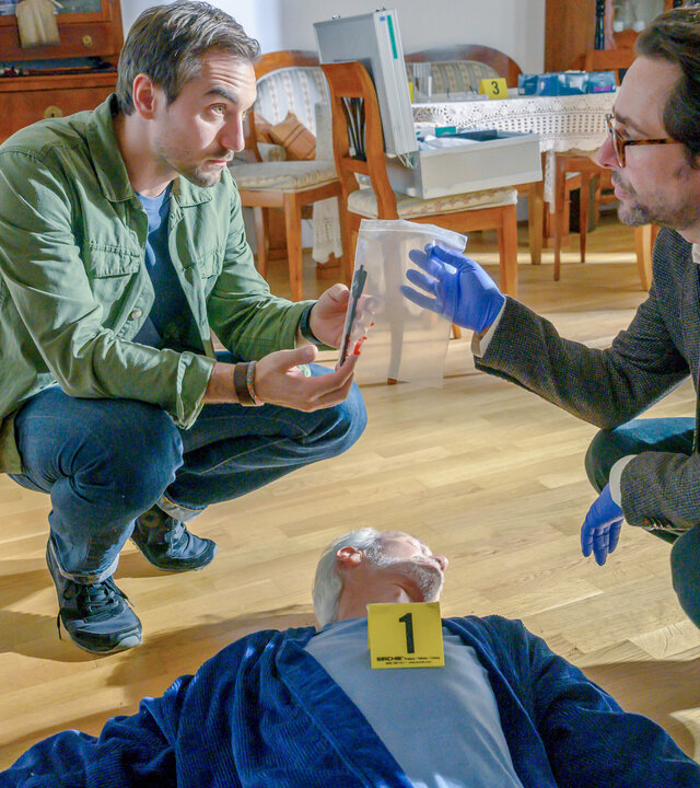 "SOKO Linz - Erlösung": Ben Halberg (Daniel Gawlowski) und Dr. Richard "Richie" Vitek (Alexander Pschill) kauern auf dem Boden über einem Mann, der dort reglos liegt. Richie übergibt Ben dabei ein durchsichtiges Tütchen, in dem sich ein länglicher Gegenstand befindet.