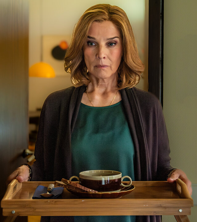 "In fremden Händen": Die elegant gekleidete Karola (Désirée Nosbusch) hält ein Essenstablett in den Händen. Ihr unergründlicher Blick geht in die Richtung, in die sie geht. Im Hintergrund gibt eine offene dunkle Holztür den Blick auf ein Esszimmer mit einer runden, orangen Hängeleuchte frei.