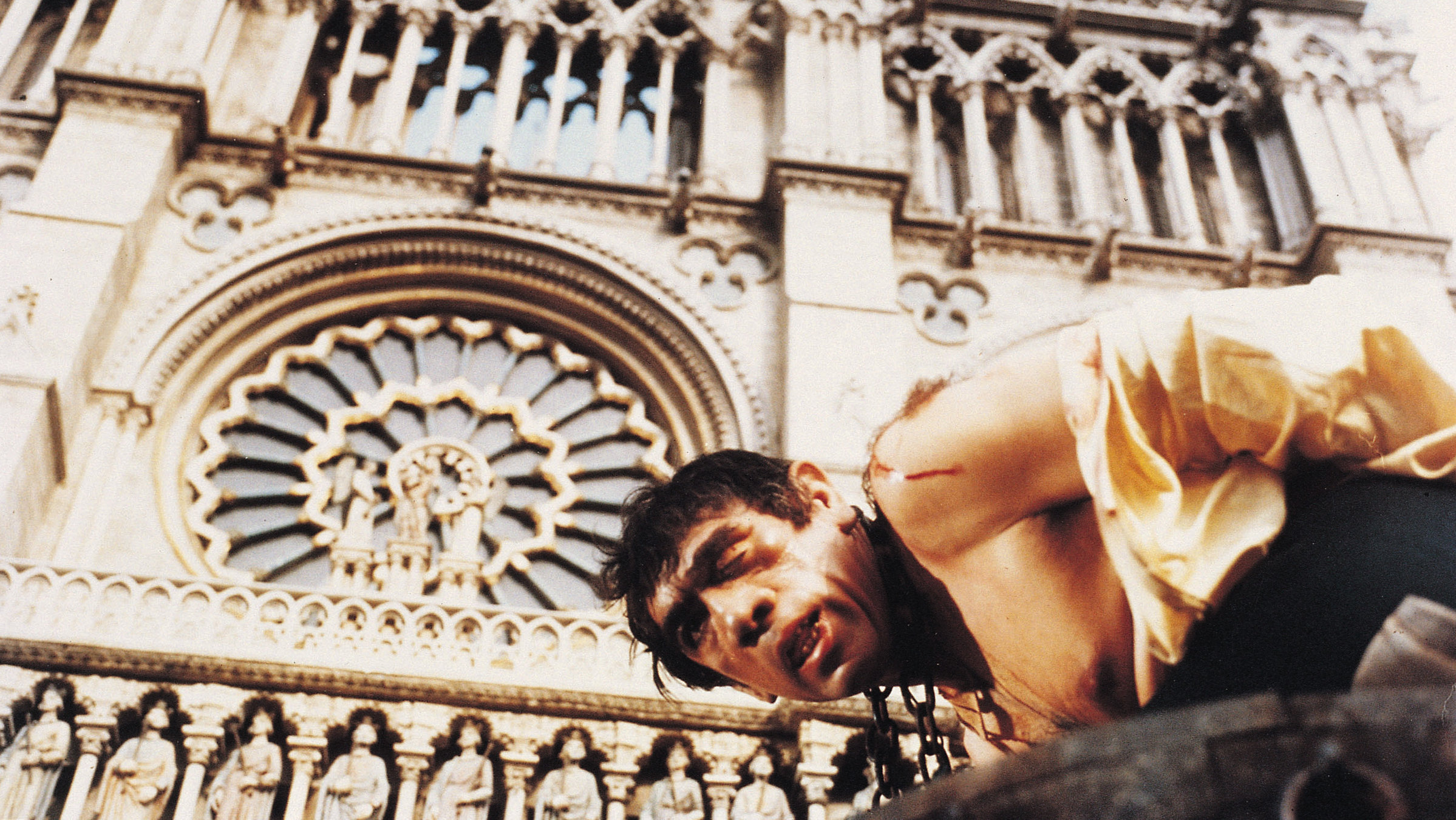 "Der Glöckner von Notre Dame": Quasimodo, ein dunkelhaariger Mann mit schiefem Gesicht und hässlichen Zähnen kniet mit einer schweren Eisenkette um den Hals vor der Kathedrale Notre Dame. Das Hemd hängt ihm in Fetzen vom Körper, er blutet an der Schulter.
