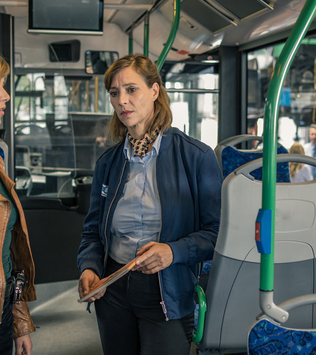 "SOKO München - Endstation": Sabine Preitl (Carin C. Tietze) steht im Gang eines Linienbusses. Sie hält eine Broschüre in der Hand und blickt ins Leere. Resi Schwaiger (Mersiha Husagic) steht links neben ihr und spricht zu ihr. Durch das Fenster des Busses sieht man eine Gruppe anderer Busfahrerdraußen stehen.