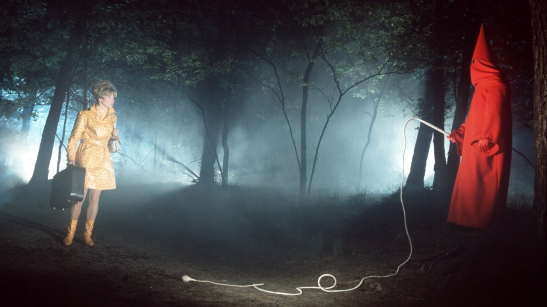 "Der Mönch mit der Peitsche": Betty Falks (Grit Boettcher) steht erschrocken in einem dunklen Wald einem Mönch mit einer Peitsche gegenüber, während sie einen großen Koffer in ihrer Hand hält.