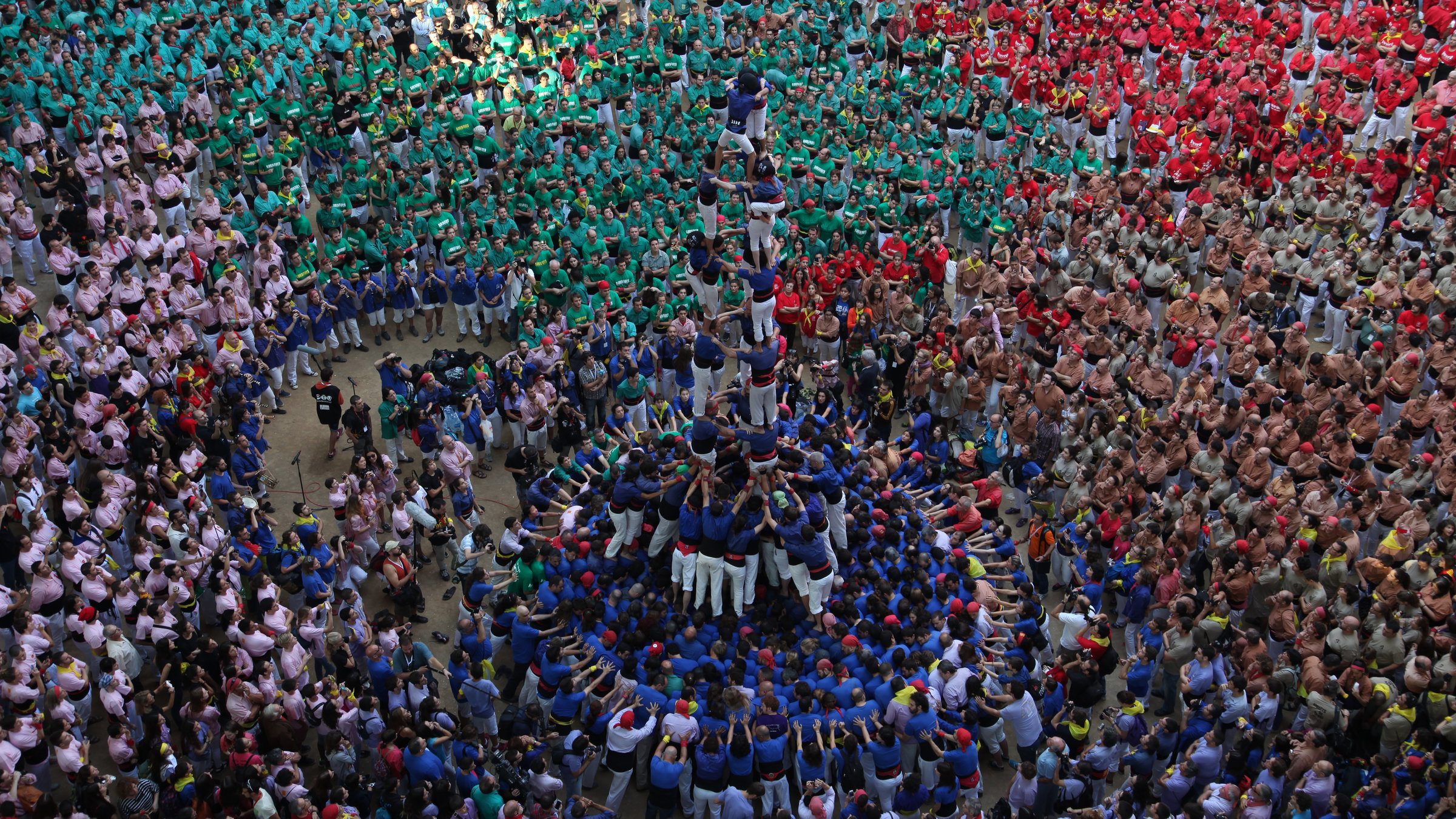 "Terra X: Fantastische Phänomene - Welt in Bewegung": Vogelperspektive - Viele Menschen in blauen T-Shirts bilden einen der Menschentürme in Vilafranca, Spanien. Um sie herum stehen bildfüllend diverse Menschengruppen mit jeweils andersfarbigen T-Shirts.
