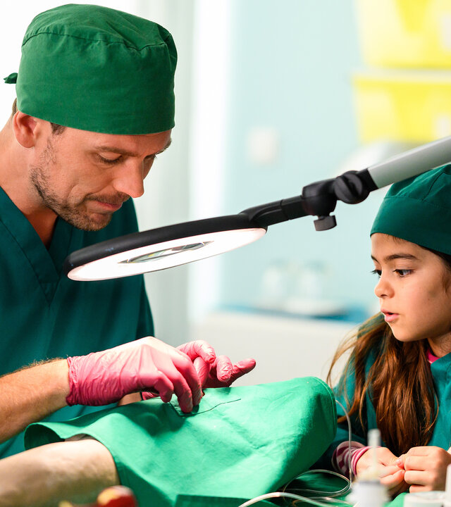 "Bettys Diagnose - Liebe hat viele Gesichter": Dr. Walden (Constantin Lücke) schaut durch eine OP-Lampe und erklärt der kleinen Jonna (Soraya Maria Efe), wie eine OP funktioniert. Im Vordergrund sind verschiedene OP-Utensilien zu erkennen.