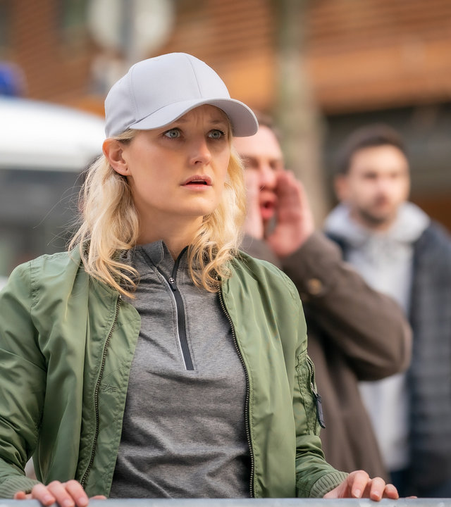 "Furia (3)": Ragna (Ine Marie Wilmann) steht an einer Straßensperrung und schaut mit geschocktem Blick in eine Richtung. Hinter ihr ist ein Polizeiauto und Demonstranten zu erkennen.