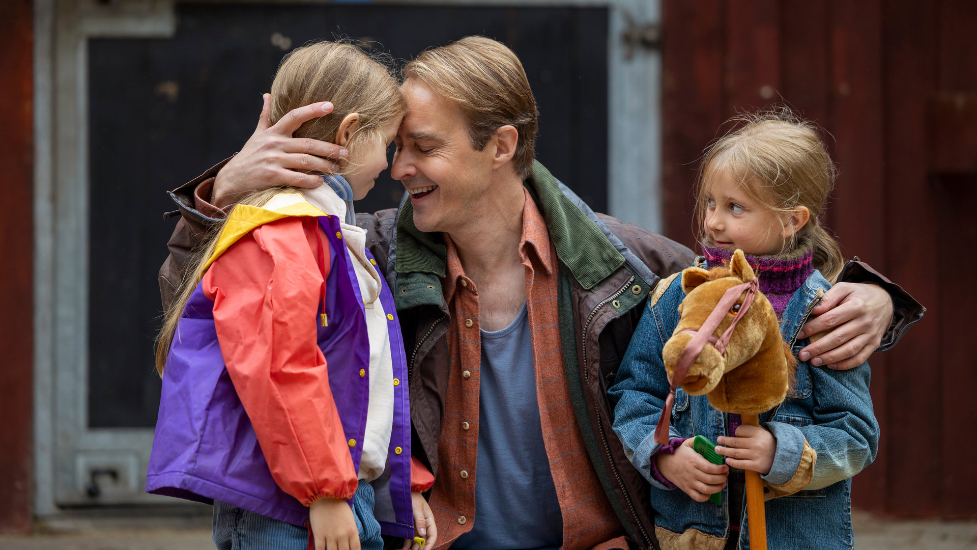 "Riding in Darkness - Aussage gegen Aussage": Tommy (Jonas Karlsson) ist in die Hocke gegangen und drückt seine beiden Töchter Victoria (Emma Comstedt) und Agnes (Lovis Bello) an sich. Im Hintergrund ist ein Haus des Reiterhofs zu sehen.