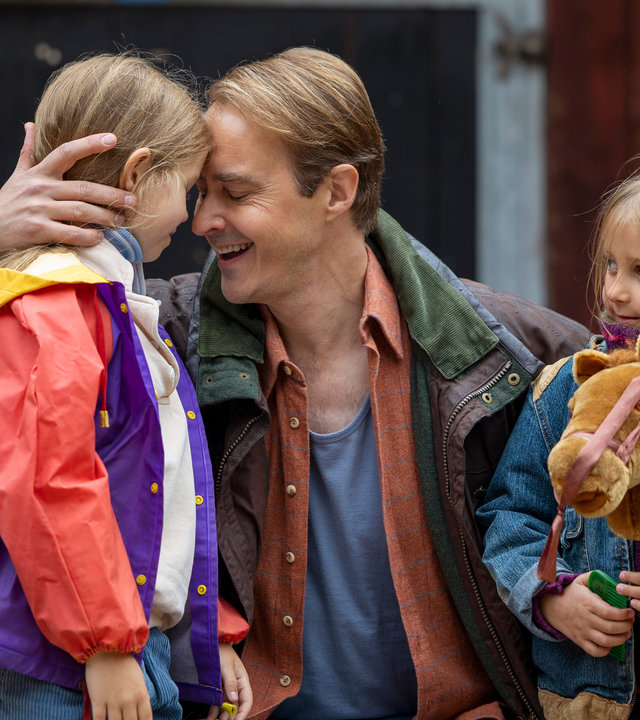 "Riding in Darkness - Aussage gegen Aussage": Tommy (Jonas Karlsson) ist in die Hocke gegangen und drückt seine beiden Töchter Victoria (Emma Comstedt) und Agnes (Lovis Bello) an sich. Im Hintergrund ist ein Haus des Reiterhofs zu sehen.