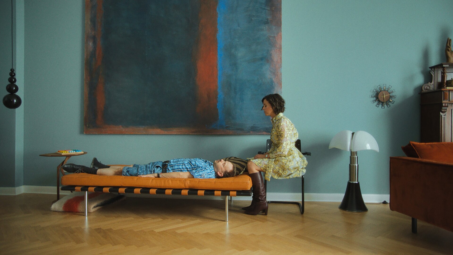 "Doppelhaushälfte - Therapie": Andi (Milan Peschel) liegt mit ausgestreckten Armen auf einer cognacfarbenen Lederliege in einem stilvoll-minimalistischen Wohnzimmer. Am Kopfende sitzt seine Therapeutin (Alice Gruia) auf einem Stuhl und beobachtet ihn, während er wie in Trance wirkt.
