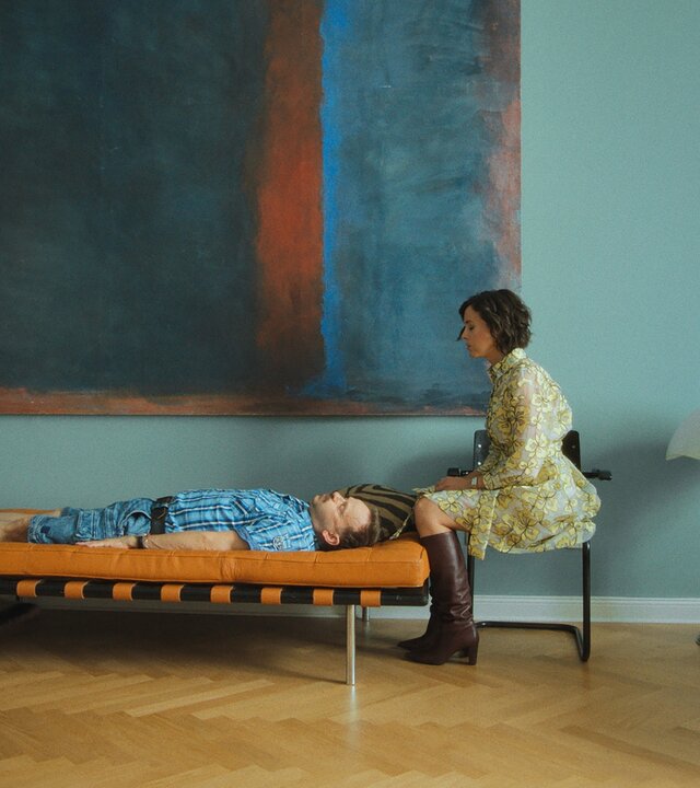 "Doppelhaushälfte - Therapie": Andi (Milan Peschel) liegt mit ausgestreckten Armen auf einer cognacfarbenen Lederliege in einem stilvoll-minimalistischen Wohnzimmer. Am Kopfende sitzt seine Therapeutin (Alice Gruia) auf einem Stuhl und beobachtet ihn, während er wie in Trance wirkt.