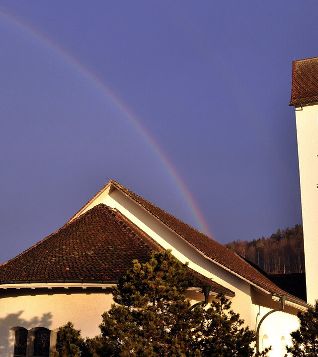 "Evangelischer Gottesdienst - Zusammen geht es besser": Vor einem tiefblauen Himmel spannt sich ein Regenbogen, der vom Dach der Kirche Gümligen aufzusteigen scheint. Der weiße Kirchturm ragt auf der einen Seite in die Höhe, davor liegt das Satteldach der massiven Kirche. Die Kirche ist weiß gestrichen und von Bäumen umgeben.