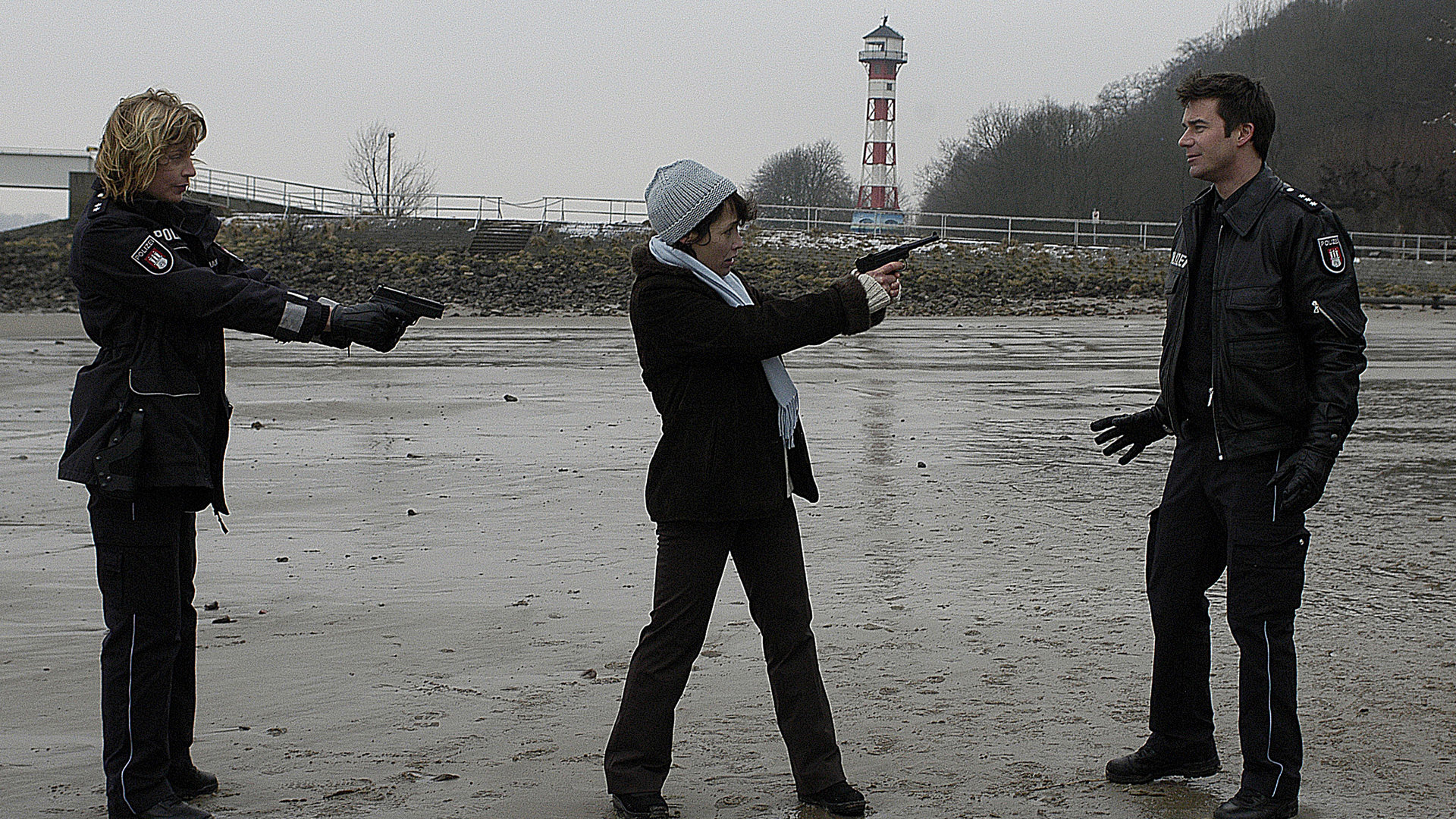 "Notruf Hafenkante - Das kalte Herz": Auf dem Wattenmeer bedroht Bettina Schmidt (Anke Sevenich) Nils (Thomas Scharff) mit einer Pistole. Melanie (Sanna Englund) steht daneben und bedroht wiederum Bettina Schmidt mit ihrer Waffe.