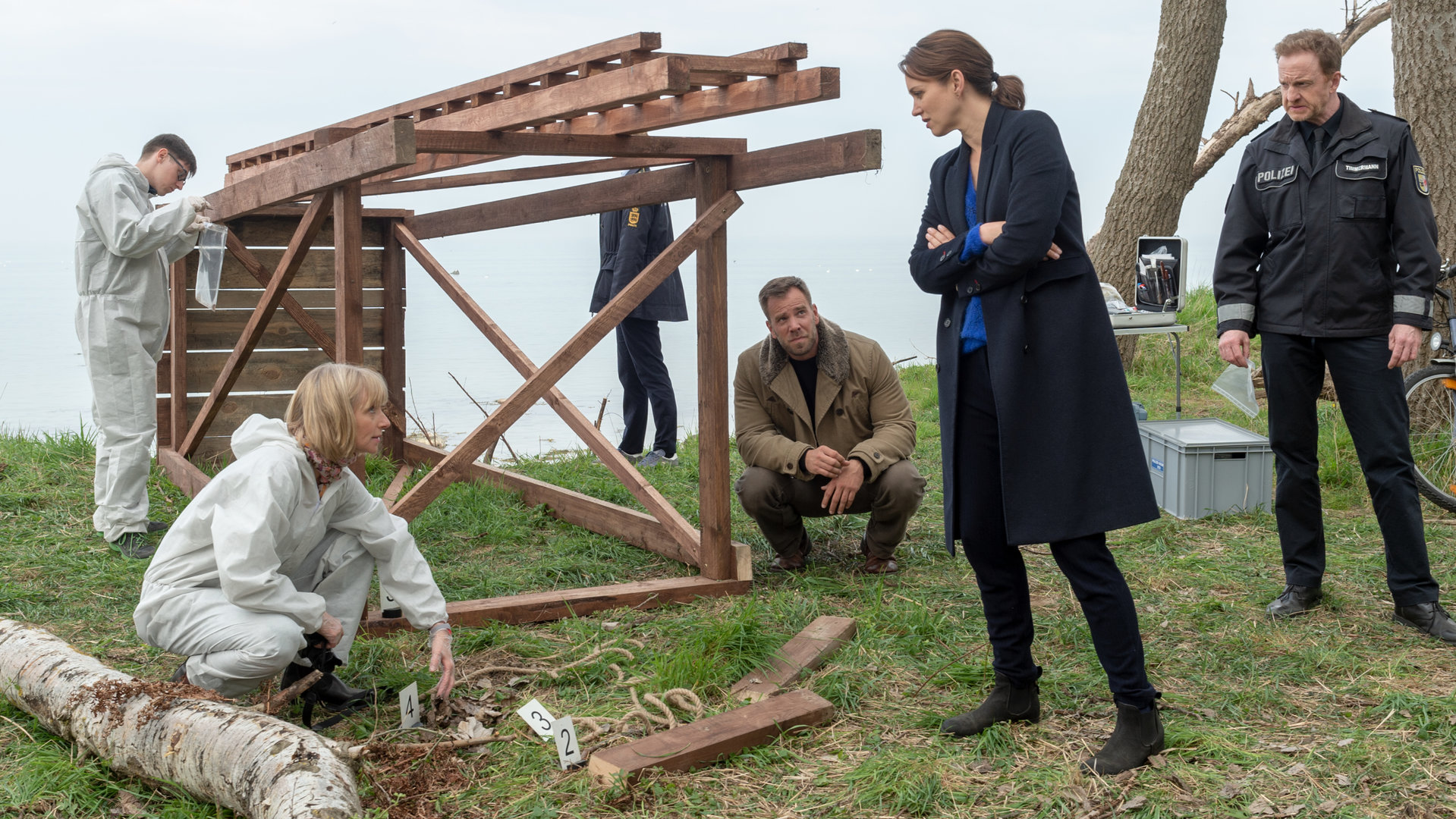 "SOKO Wismar – Tote Katze, toter Lehrer": An einem begrünten Hang in Wassernähe ist ein Hochsitz umgestürzt. Roswitha Prinzler (Silke Matthias) kniet links davon. Vor ihr liegen die angesägten Stelzen des Hochsitzes. Kommissarin Joost (Nike Fuhrmann) und Timmermann (Mathias Junge) stehen rechts vom Hochsitz, Kollege Pöhlmann (Dominic Boeer) hockt daneben. Alle begutachten die manipulierte Stelle.