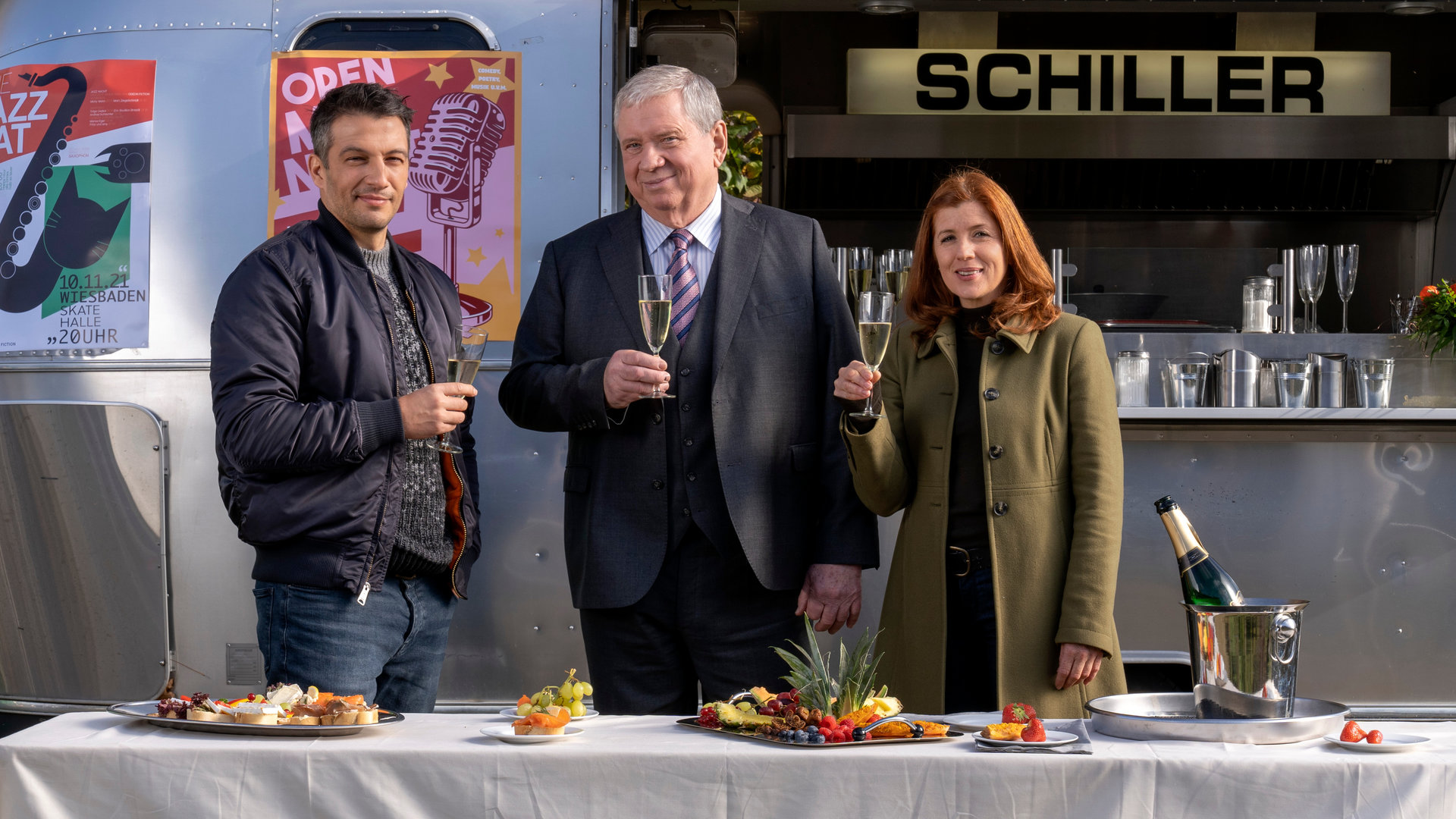 "Der Staatsanwalt - Aus der Schusslinie": Oberstaatsanwalt Bernd Reuther (Rainer Hunold), Hauptkommissarin Kerstin Klar (Fiona Coors) und Oberkommissar Max Fischer (Max Hemmersdorfer) stehen vor einem Imbisswagen und blicken zufrieden in die Kamera. Alle halten ein Sektglas in der Hand. Vor ihnen auf einem Tisch stehen einige Häppchen, eine Platte mit Obst und ein Flaschenkühler, in dem sich eine geöffnete Sektflasche befindet.