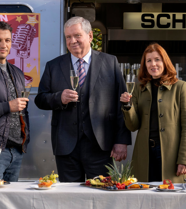 "Der Staatsanwalt - Aus der Schusslinie": Oberstaatsanwalt Bernd Reuther (Rainer Hunold), Hauptkommissarin Kerstin Klar (Fiona Coors) und Oberkommissar Max Fischer (Max Hemmersdorfer) stehen vor einem Imbisswagen und blicken zufrieden in die Kamera. Alle halten ein Sektglas in der Hand. Vor ihnen auf einem Tisch stehen einige Häppchen, eine Platte mit Obst und ein Flaschenkühler, in dem sich eine geöffnete Sektflasche befindet.