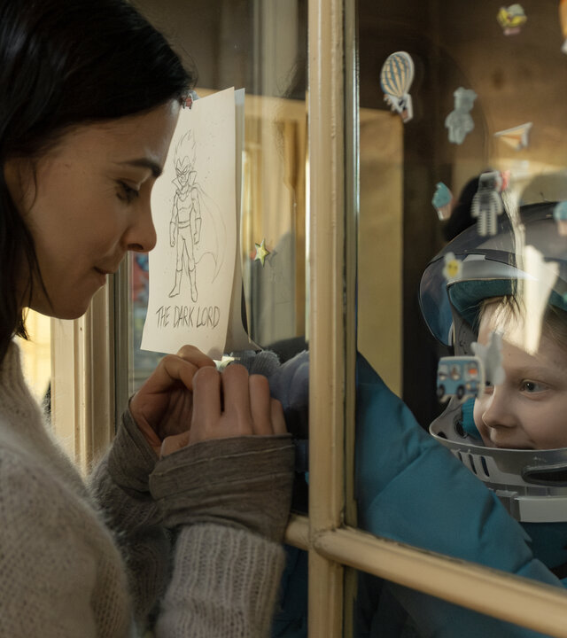 "Serial Cleaner - Kein Zurück": Teri Marquard (Aylin Tezel) und ihre schwerkranke Tochter Luca (Hanna Heckt) befinden sich in einem Raum und sind durch eine Glasscheibe getrennt. Teri hat ihre Hände zu einem Herz geformt und hält sie für ihre Tochter an die Glasscheibe. Luca trägt Schutzkleidung und schaut lächelnd zu ihrer Mutter.