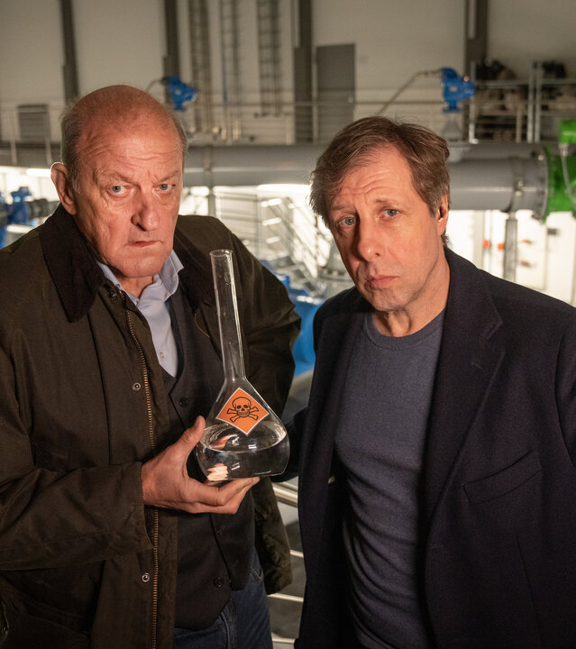 "Wilsberg - Mit allen Wassern gewaschen": In einer Fabrikhalle stehen Wilsberg (Leonard Lansink) und Ekki (Oliver Korittke) nebeneinander und schauen ernst in die Kamera. Wilsberg hält einen Messkolben in der Hand, in welchem sich Wasser befindet und der mit einem Totenkopf-Warnaufkleber versehen ist.