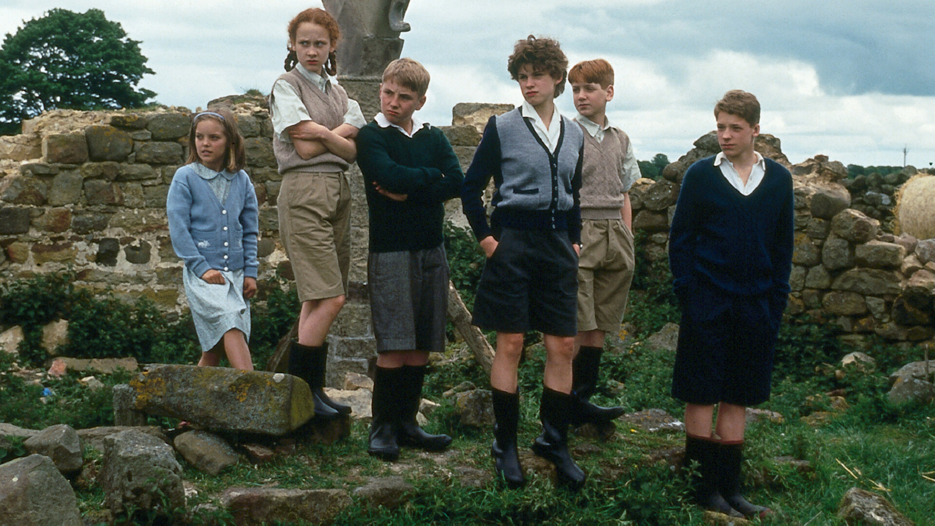"Fünf Freunde und das Burgverlies": Anne (Laura Petela), Harriet (Leanne Rowe), Dick (Paul Child), George (Jemima Rooper), Harry (Greg Bradley) und Julian (Marco Williamson) stehen in Gummistiefeln auf einer Wiese vor einer Steinmauer und blicken kritisch zur Seite.