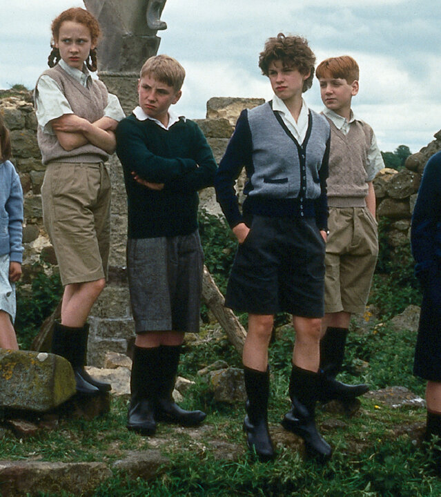 "Fünf Freunde und das Burgverlies": Anne (Laura Petela), Harriet (Leanne Rowe), Dick (Paul Child), George (Jemima Rooper), Harry (Greg Bradley) und Julian (Marco Williamson) stehen in Gummistiefeln auf einer Wiese vor einer Steinmauer und blicken kritisch zur Seite.