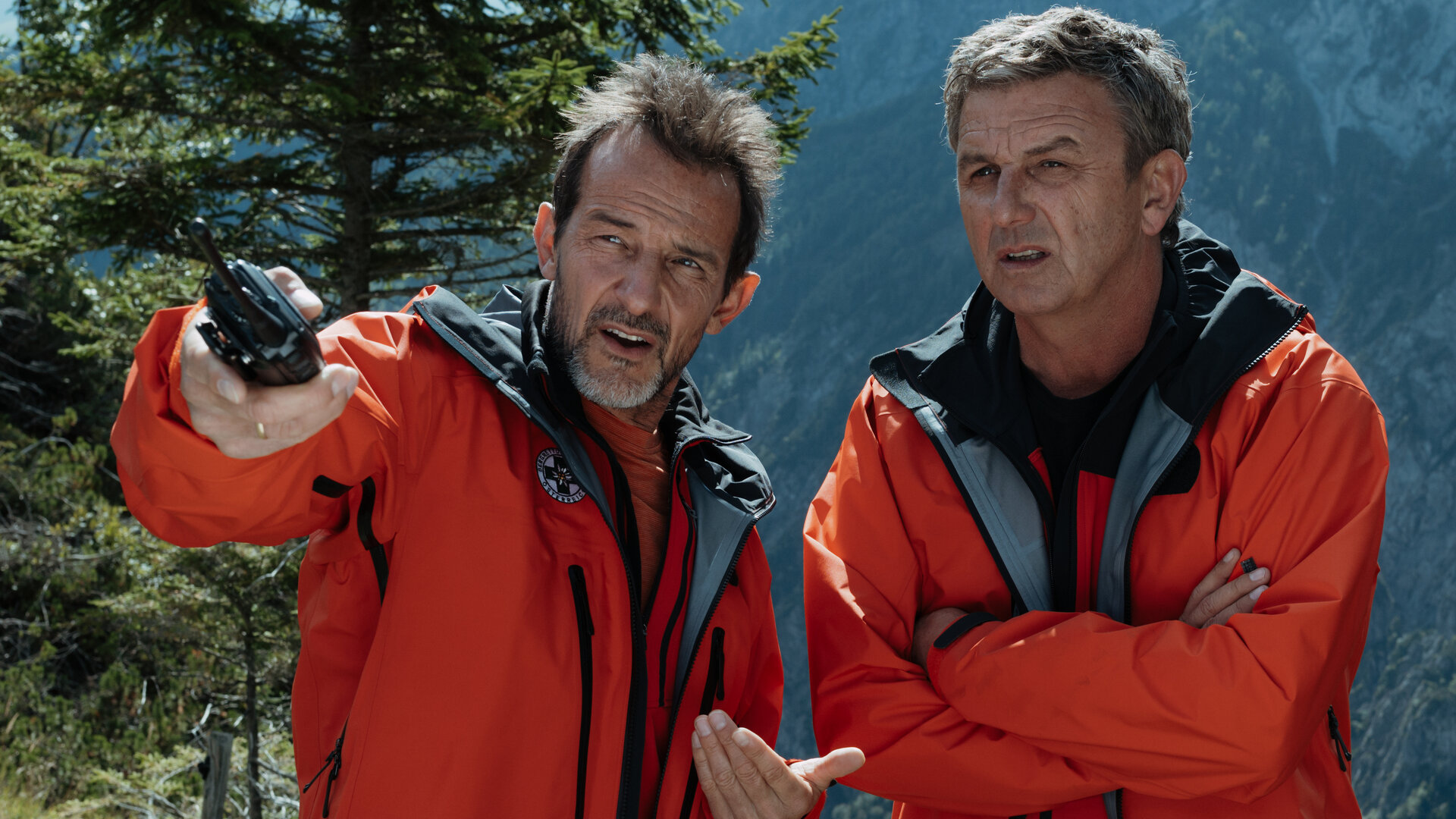 "Der Bergdoktor: Am Anschlag": In den Bergen: Moritz Stölzer (Hans-Werner Meyer) und Dr. Martin Gruber (Hans Sigl) stehen nebeneinander. Beide tragen die roten Jacken der Bergrettung. Moritz hält ein Walkie-Talkie in der Hand, mit der er auf etwas außerhalb des Bildrands zeigt. Dr. Martin Gruber hält die Arme vor der Brust verstränkt und blickt kritisch in diese Richtung.