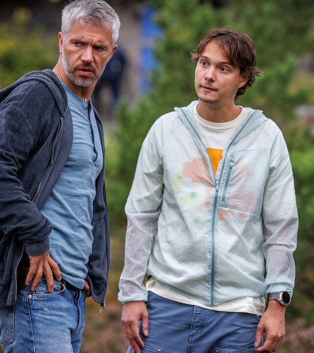 "Mord im Mittsommer - Nikki & Evelina": Alexander (Nicolai Cleve Broch) und Welpe (Julius Fleischanderl) stehen im Freien nebeneinander. Alexander wirkt skeptisch. Welpe blickt in seine Richtung.
