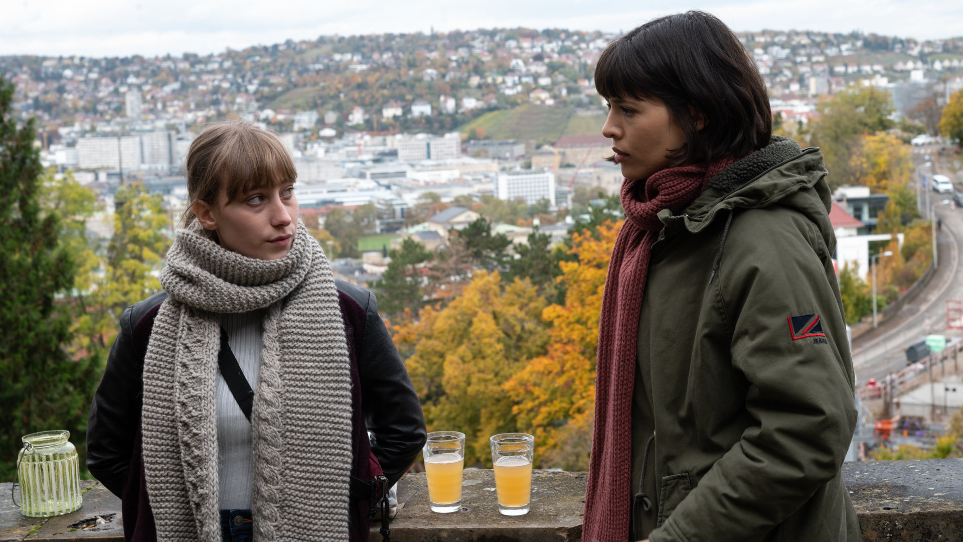 "SOKO Stuttgart – Wohnungslos": Kommissarin Lea Gomez (Jasmin Gassmann) und Emily Friedl (Juliane Schütze) stehen im Freien zusammen. Auf einer Mauer haben sie ihre Getränke abgestellt. Im Hintergrund sieht man in den Stuttgarter Stadtkessel. Die beiden Frauen schauen sich ernst an.