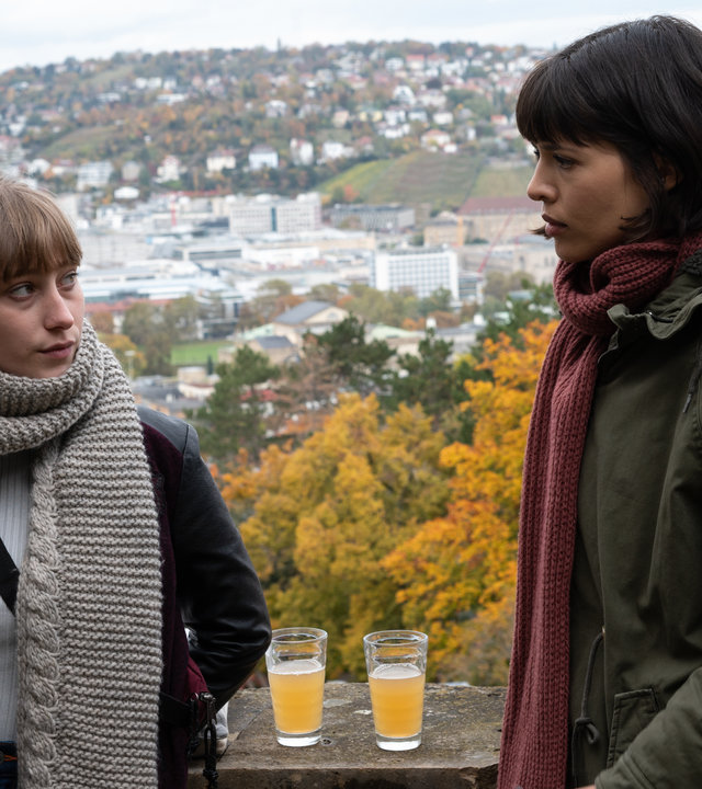 "SOKO Stuttgart – Wohnungslos": Kommissarin Lea Gomez (Jasmin Gassmann) und Emily Friedl (Juliane Schütze) stehen im Freien zusammen. Auf einer Mauer haben sie ihre Getränke abgestellt. Im Hintergrund sieht man in den Stuttgarter Stadtkessel. Die beiden Frauen schauen sich ernst an.
