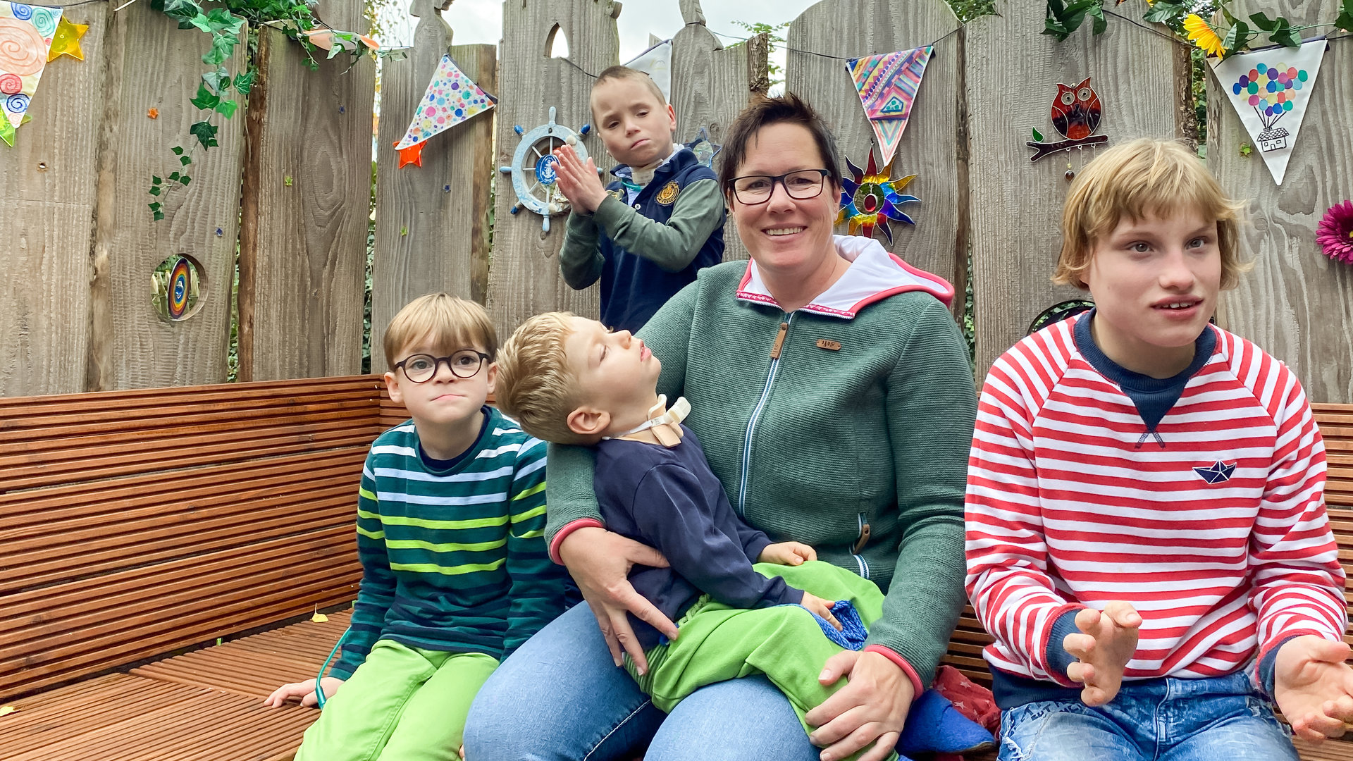 "37° Mama Held - Eine Pflegemutter kämpft für ihre Kinder": Eine Frau mittleren Alters mit kurzem dunklen Haar sitzt inmitten ihrer vier Pflegekinder auf einer Bank im Freien.
