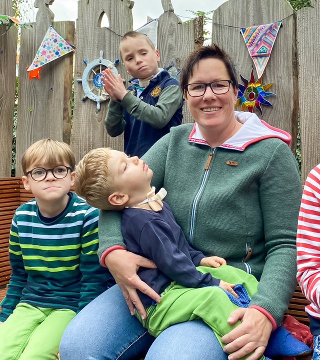 "37° Mama Held - Eine Pflegemutter kämpft für ihre Kinder": Eine Frau mittleren Alters mit kurzem dunklen Haar sitzt inmitten ihrer vier Pflegekinder auf einer Bank im Freien.
