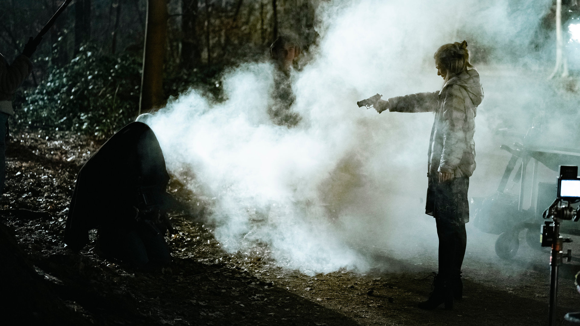 "SOKO Köln - Angriff der Schatten": Maja Kersten (Alina Bergmann) steht seitlich zur Kamera in einem düsteren Wald und zielt mit einer Pistole auf eine Person (Komparse), die auf dem Boden kauert. Die Person ist auf Grund der Lichtverhältnisse und einer dunklen Kapuze, die über den Kopf gezogen wurde, nicht zu erkennen. Links im Bild sieht man ein Stabmikrofon, das ins Bild gehalten wird.  Maja steht in einem dichten Nebel, der durch eine Maschine produziert wurde. Rechts im Bild sieht man eine Kamera mit leuchtendem Bildschirm auf eine Stativ stehen.
