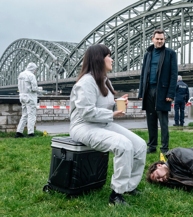 "SOKO Köln - Freiheit": Amira Mahdi (Aylin Ravanyar) und Jonas Fischer (Lukas Piloty) stehen auf einer Wiese am Rhein vor einer großen stählernen Brücke und sprechen mit Dr. Pilar Westphal (Yulia Yáñez Schmidt), die auf einem schwarzen Rollkoffer sitzt und einen Kaffee in der Hand hält. Sie trägt einen weißen Overall. Zwischen ihnen liegt der tote Jan Meyers (Anselm Ferdinand Bresgott) seitlich auf der Wiese mit einer Wunde am Kopf. Dahinter steht ein Mitarbeiter (Komparse) der Spurensicherung an einer Mauer, daneben vor einem Absperrband ein uniformierter Polizist (Komparse). In einiger Entfernung ragt im Hintergrund der Kölner Dom empor.