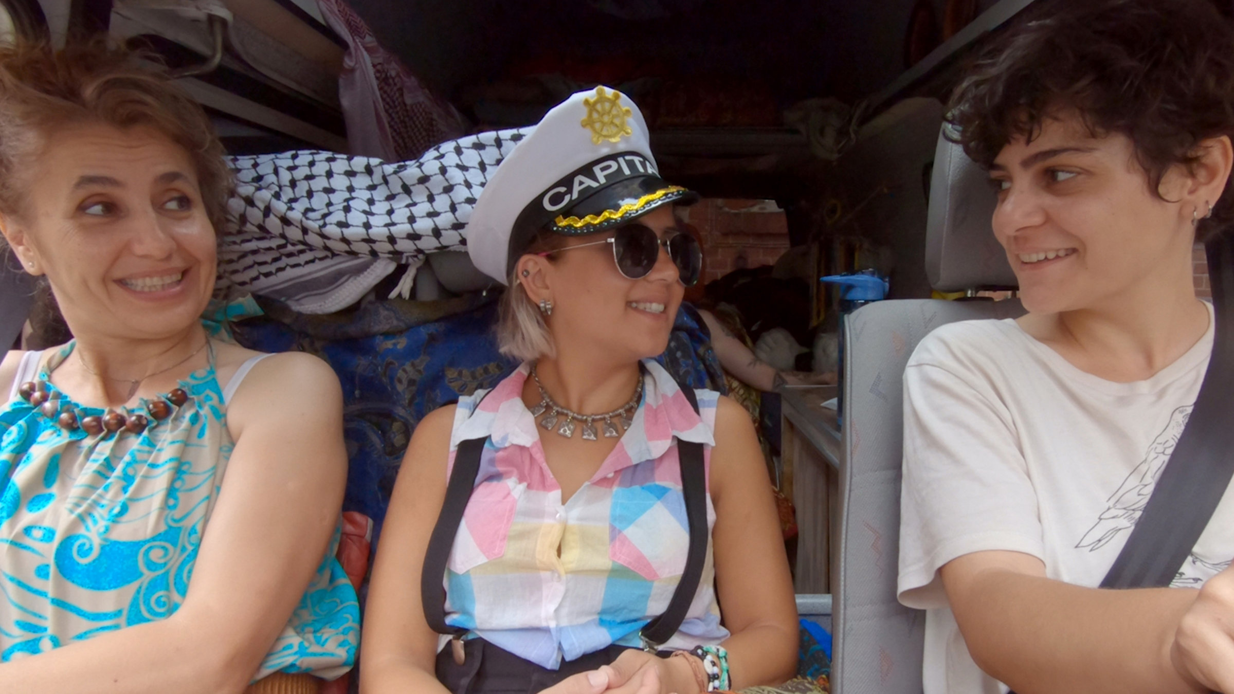 "Die Sonne ist überall gelb": Rawa, Rahaf und Afraa sitzen nebeneinander im Campingbus und lächeln sich gegenseitig an. Rahaf trägt eine Kapitänsmütze.