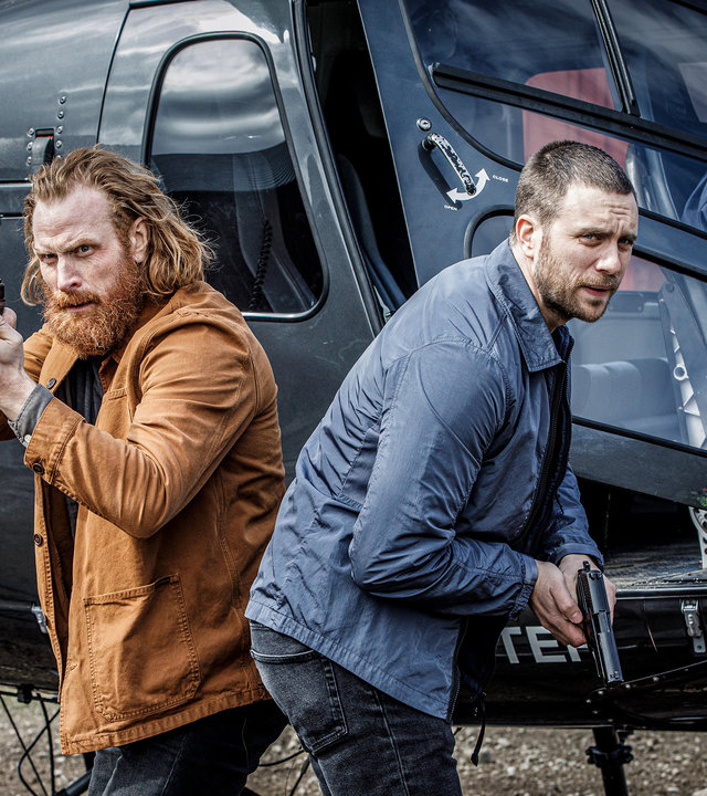 "Kommissar Beck: Ein neues Leben": Steinar Hovland (Kristofer Hivju) und Josef Eriksson (Martin Wallström) stehen mit gezogenen Waffen Rücken an Rücken vor einem Hubschrauber.